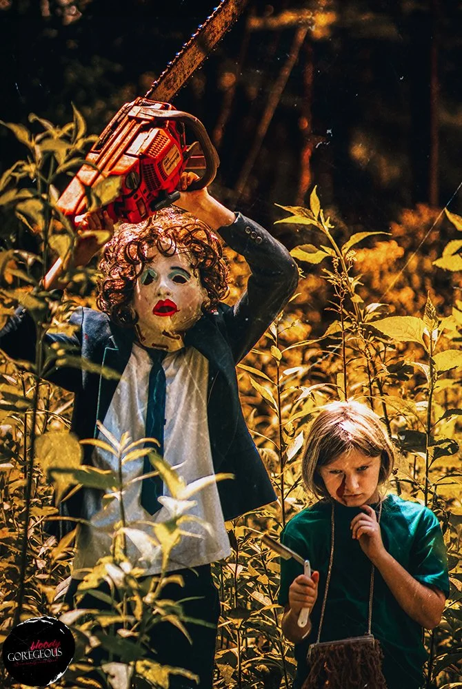 A person in a mask with red lipstick, holding a chainsaw above their head, standing among yellow plants, with a young girl nearby holding a stick and looking concerned.