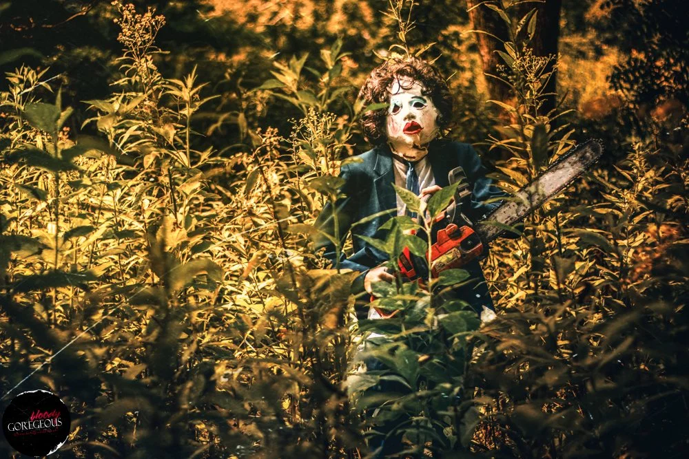 A person wearing a clown mask and dark clothing holding a chainsaw, standing amidst tall bushes or plants in a wooded area at dusk or night.