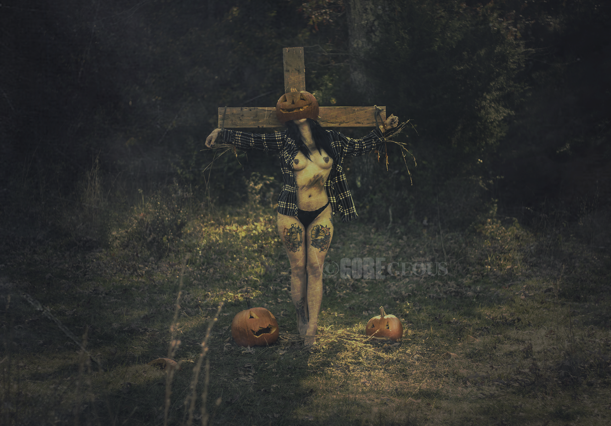 A scarecrow with a carved pumpkin head featuring a jack-o'-lantern face, wearing a plaid shirt, standing in a dark wooded area. It has tattoos on its body, arms stretched out, with a wooden crossbeam on its shoulders, and is surrounded by three carve