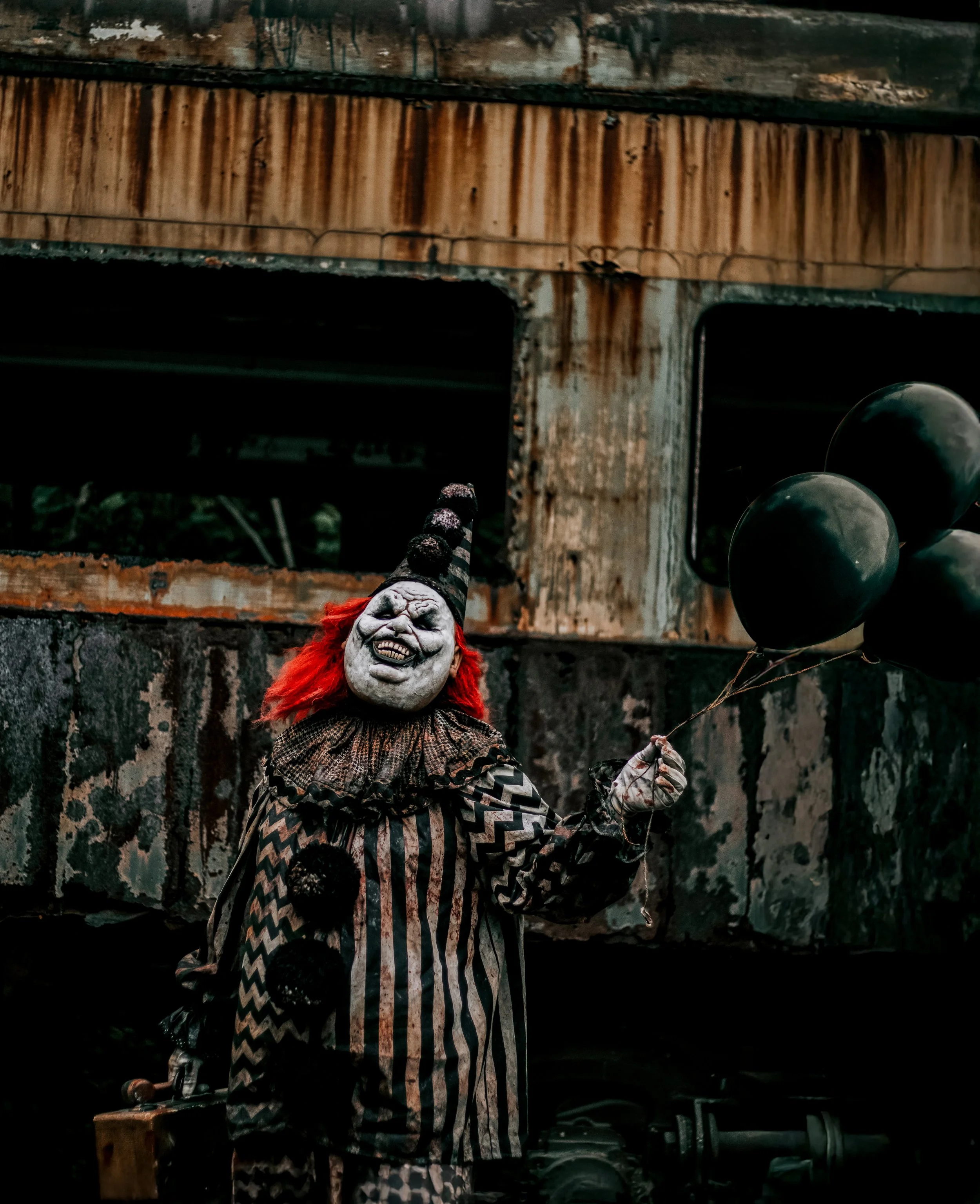 A clown with white face paint, red hair, and a striped costume holding black balloons.