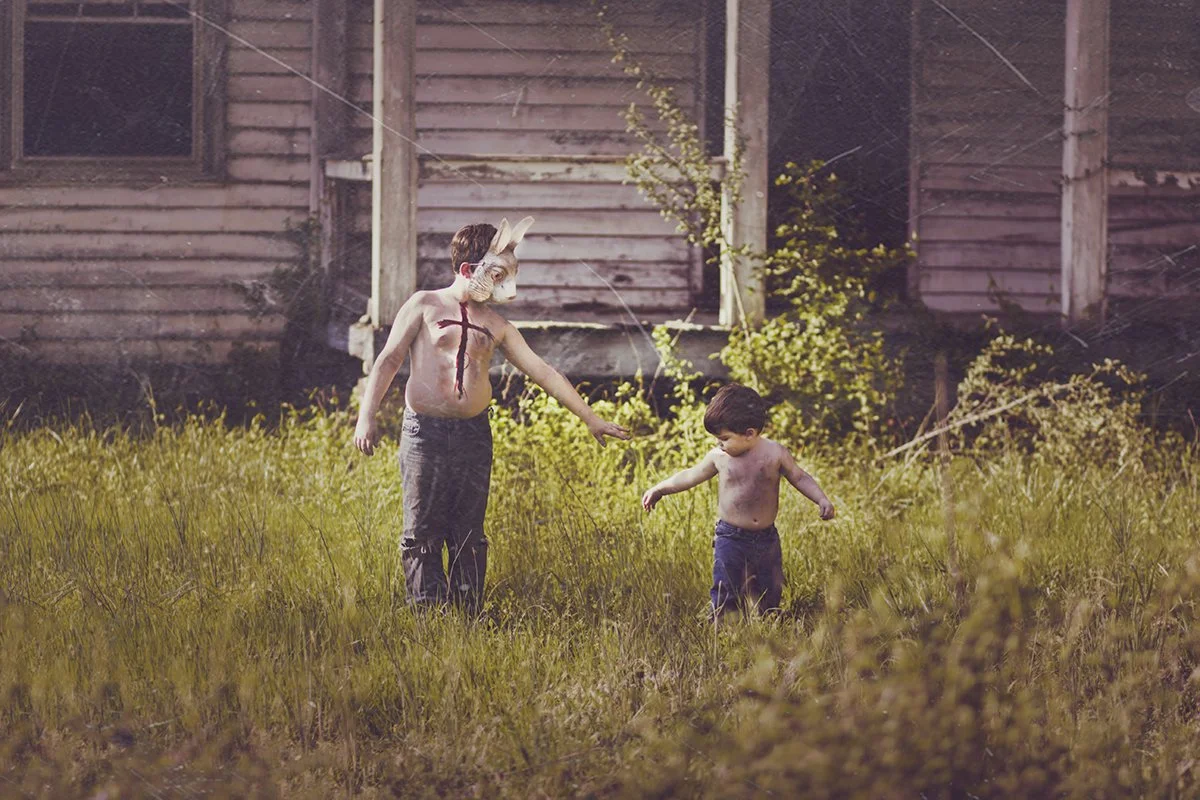 A person wearing a rabbit mask and body paint is reaching out to a young boy in a grassy yard of an old house.