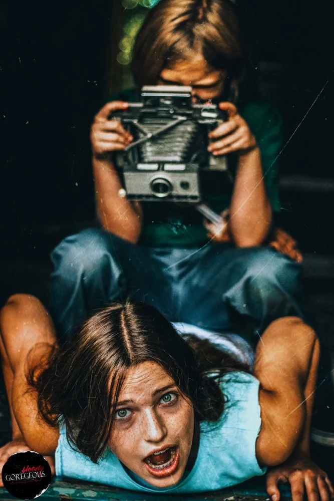 A woman with short brown hair and freckles lying face down with her arms bent, mouth open, and wide eyes. A young boy with brown hair is sitting behind her, holding a camera and pointing it towards the viewer. The scene appears intense and dramatic.