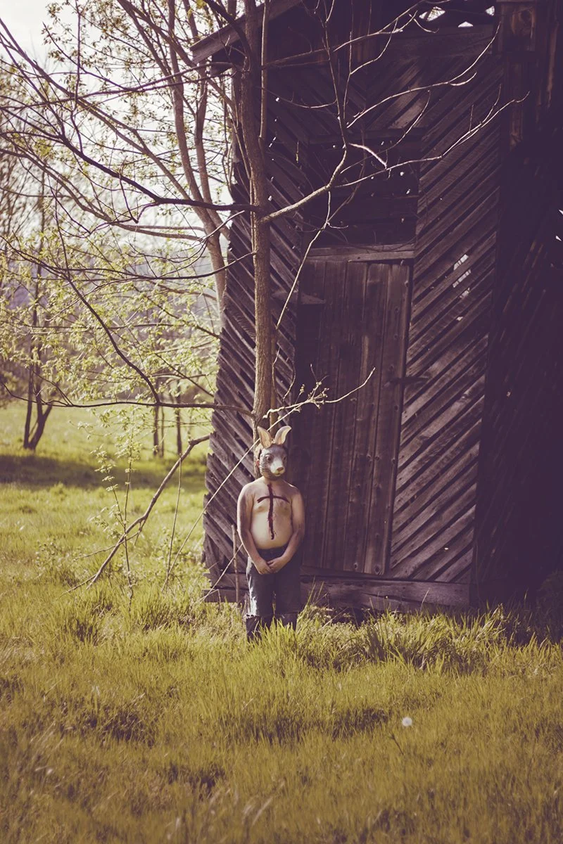A humanoid figure with a rabbit mask, no shirt, stands in a grassy area near a wooden structure. The person has a painted black cross on their chest and is holding their hands together in front. A tree with thin branches is nearby.