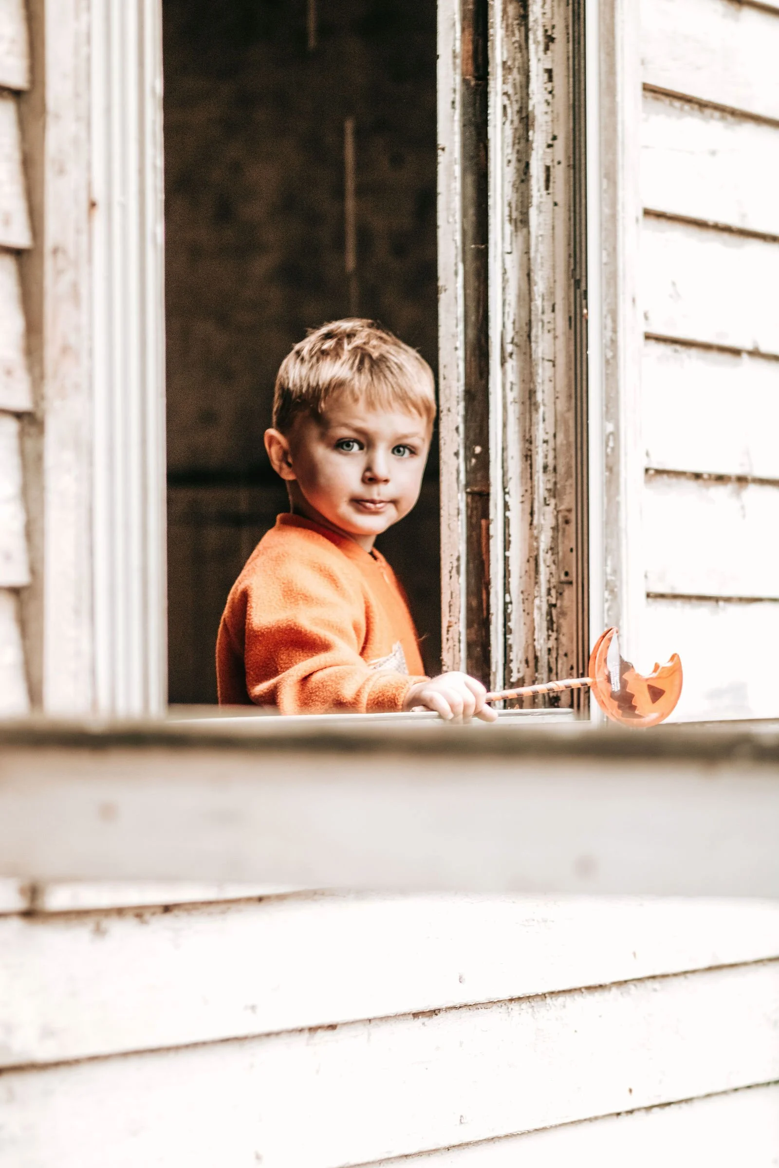A young boy with light brown hair and blue eyes wearing an orange sweater, standing at a window with peeling wood siding, holding a small plastic pumpkin-shaped Halloween basket.