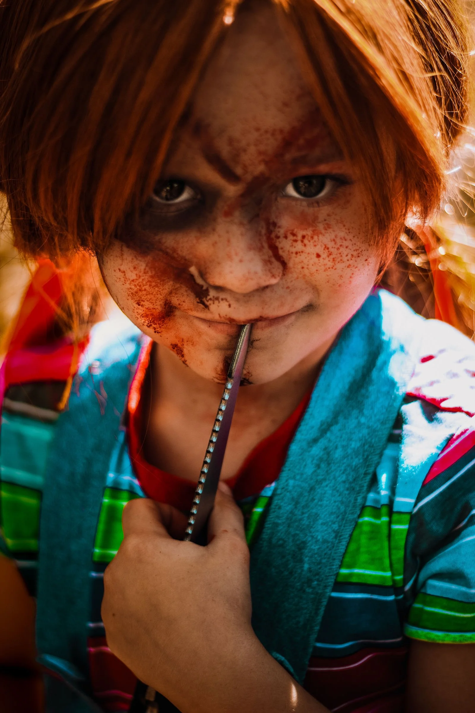Close-up of a young person with red hair, freckles, holding a spoon near their lips, wearing a colorful striped shirt and a blue vest, with sunlight and blurred outdoor background.