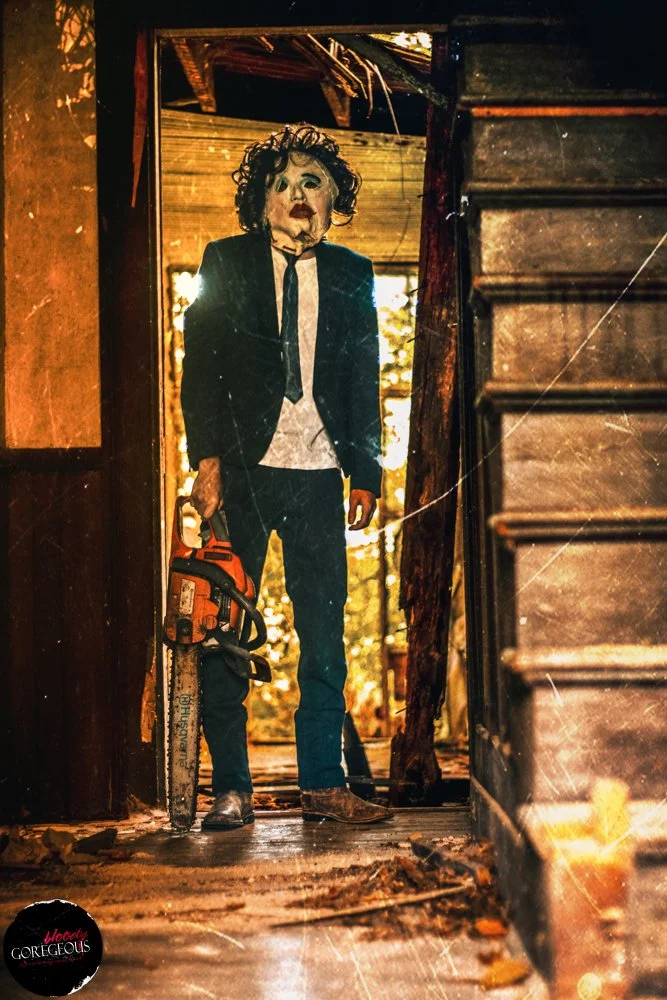 A person wearing a Michael Myers mask from the Halloween movies, dressed in a black suit with a white shirt and black tie, stands in a doorway holding a chainsaw, inside an abandoned, burned house with broken walls and debris on the floor.