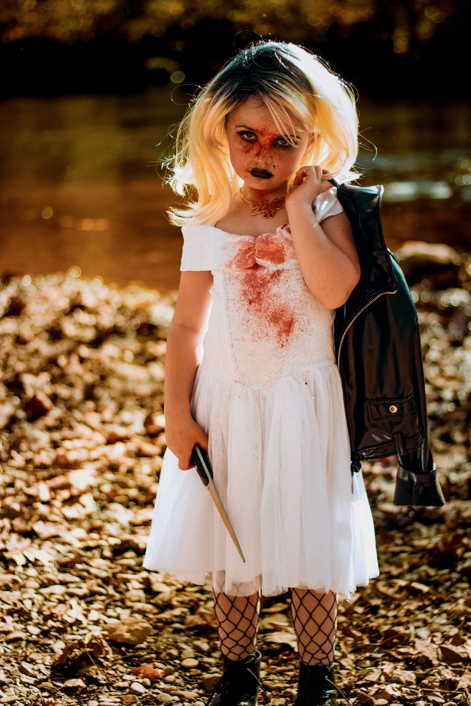 A young girl wearing a white dress with bloodstains, fishnet stockings, and black boots, standing outdoors on autumn leaves, holding a small object in her left hand, and adjusting a black leather jacket over her shoulder. Her face has dark makeup and