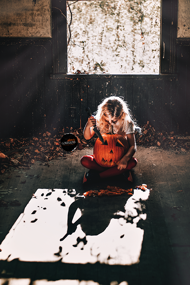 A young girl with blonde hair, wearing a white shirt and red tights, sits cross-legged on the floor of a dark room, carving a face into a large pumpkin with a knife. Sunlight streams through a window behind her, casting shadows of leaves on the floor