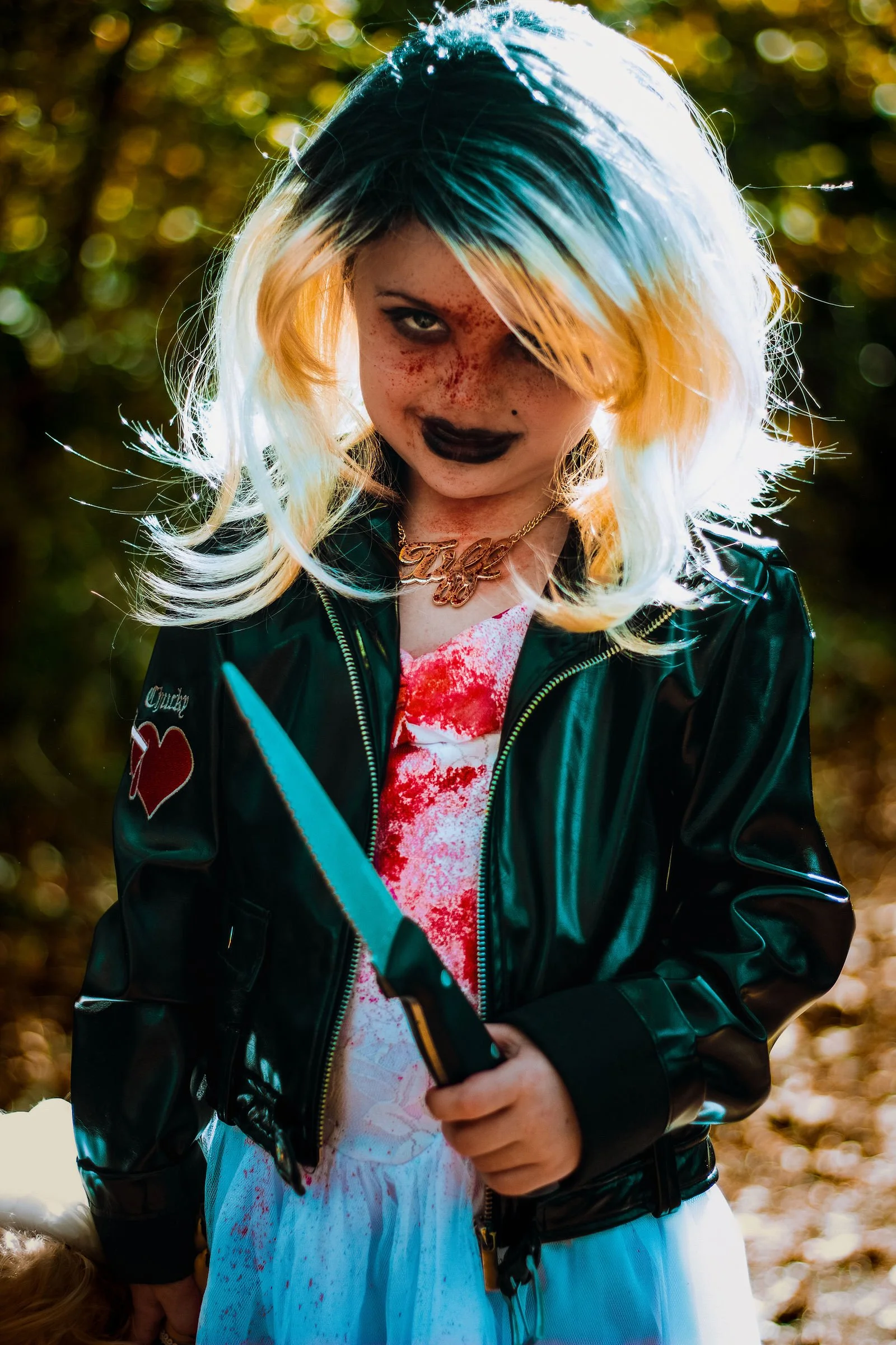 A woman with blonde and blue hair, dark makeup, and freckles is wearing a black leather jacket with patches, a necklace with a gold nameplate, and a white dress with red splatters. She is holding a phone in her right hand, standing outdoors with a ba
