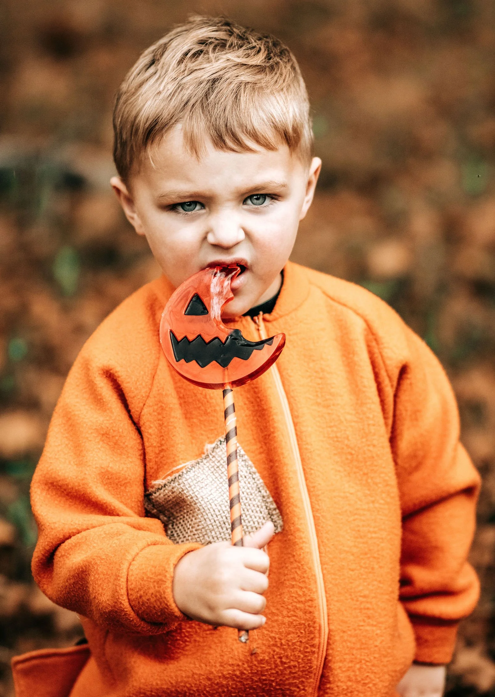 A young boy with blond hair and blue eyes wearing an orange zip-up jacket, holding a Halloween-themed candy lollipop shaped like a jack-o'-lantern.