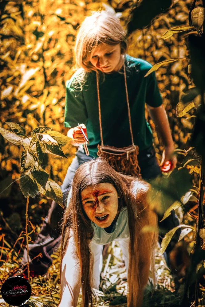 Two young girls are in a dense, sunlit forest. One girl with long hair is crawling on the ground, looking distressed and covered in face paint or makeup. The other girl with short blonde hair is standing behind her, holding a stick and appearing to b