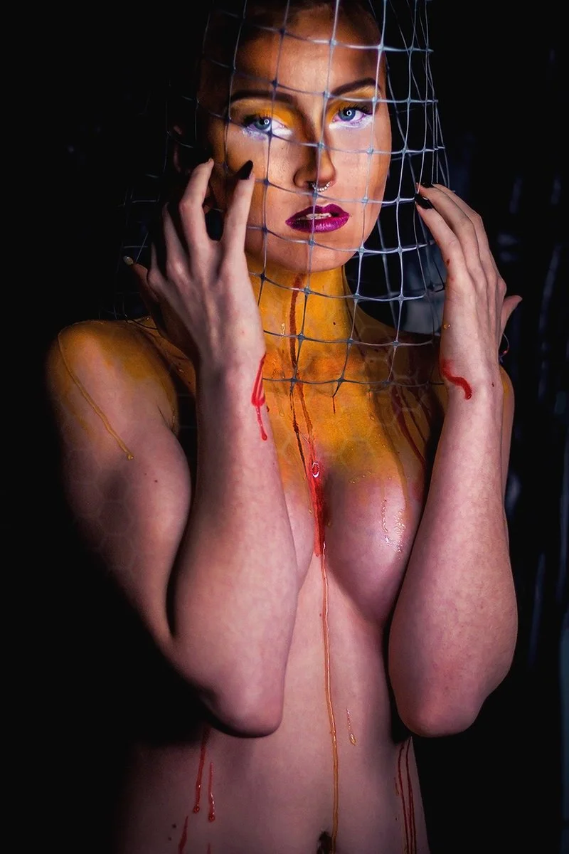 A woman with bright blue eyes and dark lipstick behind a web-like net, with colored paint and liquid dripping on her skin and face, holding the net with both hands.