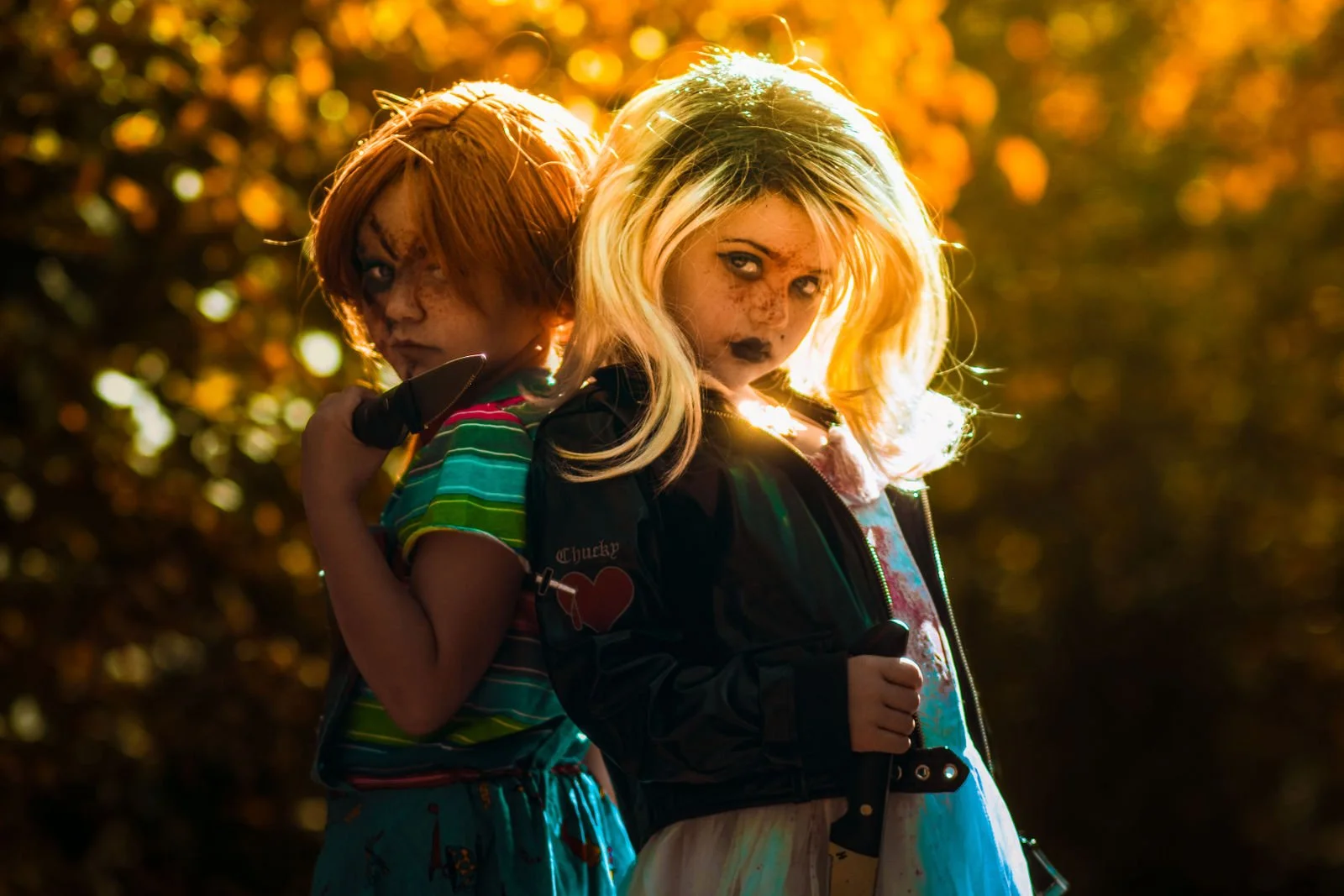Two girls dressed as horror characters with zombie makeup and costumes, holding knives, standing outdoors in front of a blurred, sunlit background.