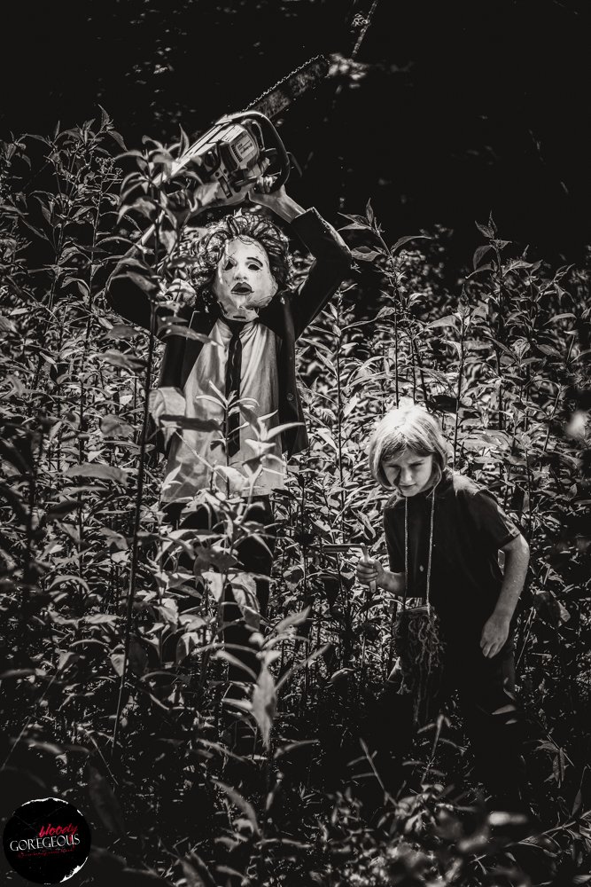 A scarecrow wearing a mask with a dress and tie holding a chainsaw, and a young girl with short hair and dark clothing holding an object, surrounded by tall plants or weeds in a field.