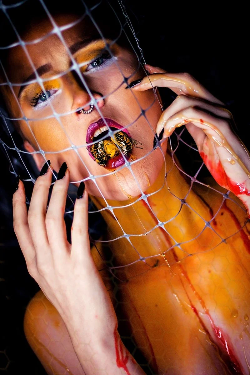 A person with artistic makeup and a septum piercing behind chicken wire, holding their hand near their face, with blood and honey on their hands and arm, and a bee on their open mouth inside the wire.