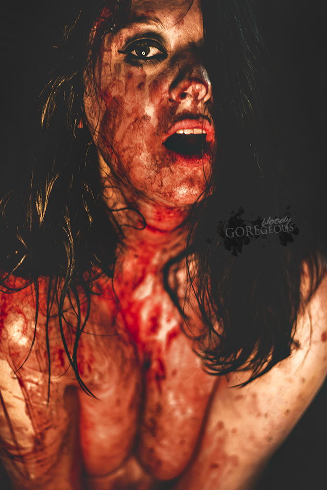 A woman with a bloody and injured face and upper body, looking distressed with her mouth open, dark hair, and a dark background.