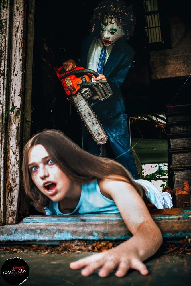 A person in a clown mask wearing a suit standing behind a woman with long brown hair, who is lying on the ground and looking scared. The clown is holding a chainsaw, and the scene appears to be staged or from a horror setup.