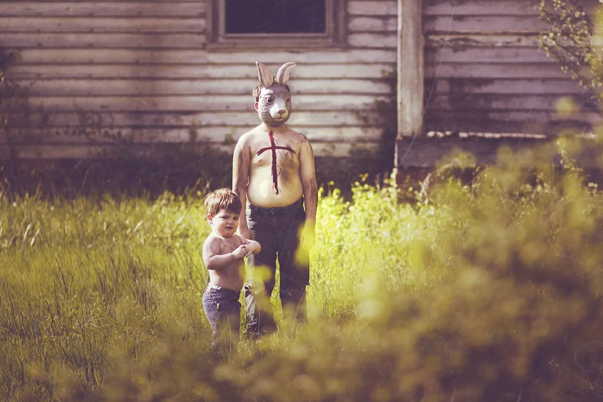 A person with a bunny mask and painted body stands in a grassy yard with a small boy, both looking at the camera, in front of a weathered wooden house.