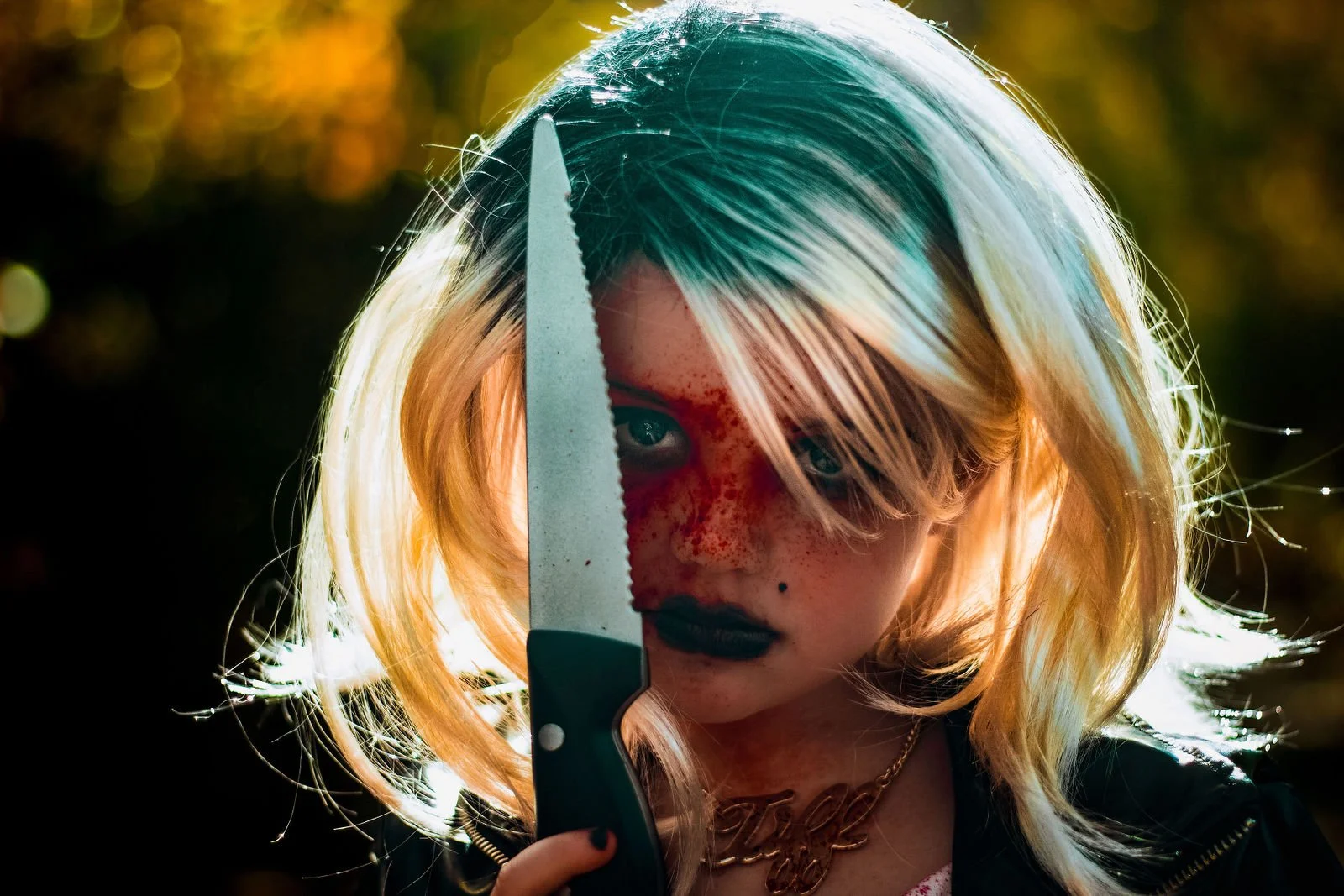 A young woman with blonde hair and freckles holding a large saw knife, with the blade partly obscuring her face. She has dark lipstick and a chain necklace, with a blurred outdoor background.