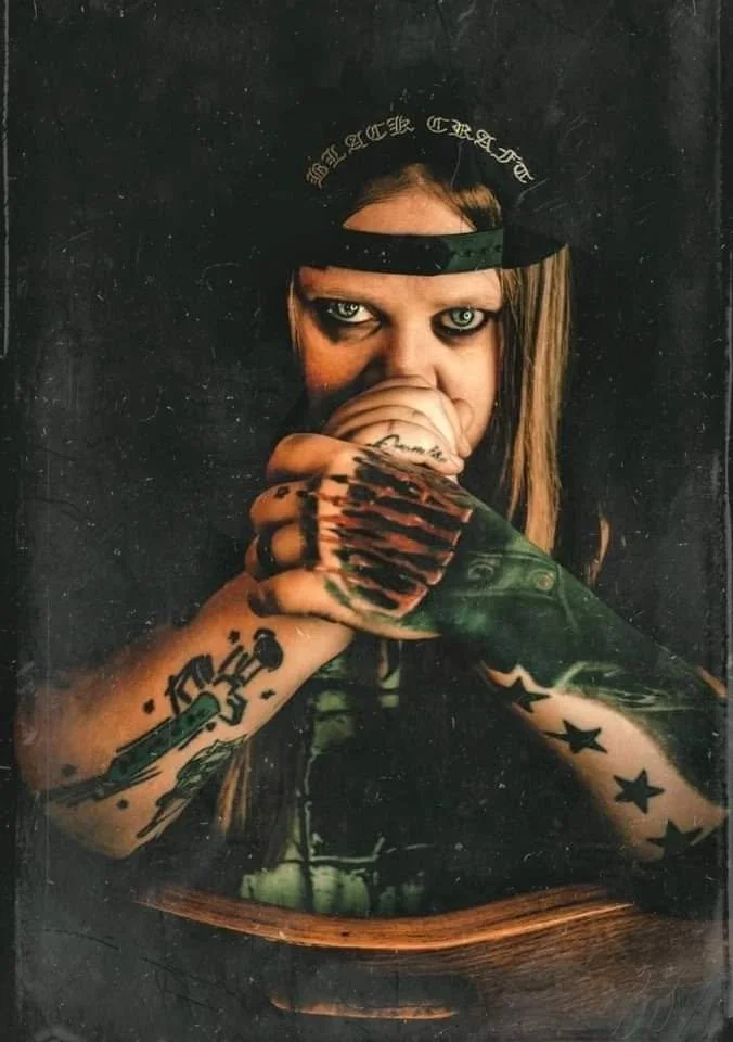 A woman with tattoos on her arms, wearing a black cap backwards, looking directly at the camera with intense eyes, holding her hand up to her face, and dressed in dark clothing against a black background.