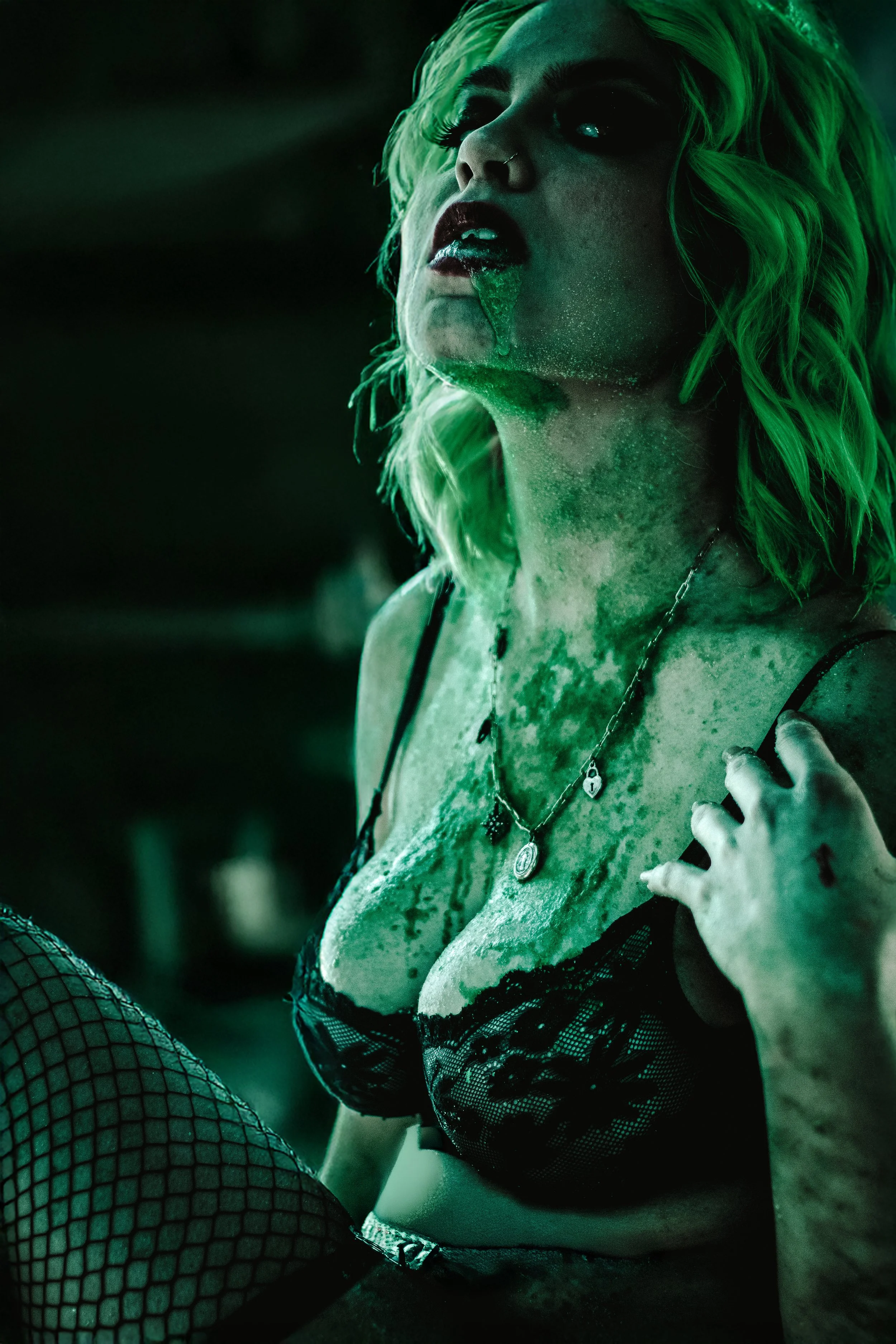 A woman with green hair and dark makeup wears fishnet stockings and black lace lingerie, with jewelry, posing with her hand on her chest against a dark background.