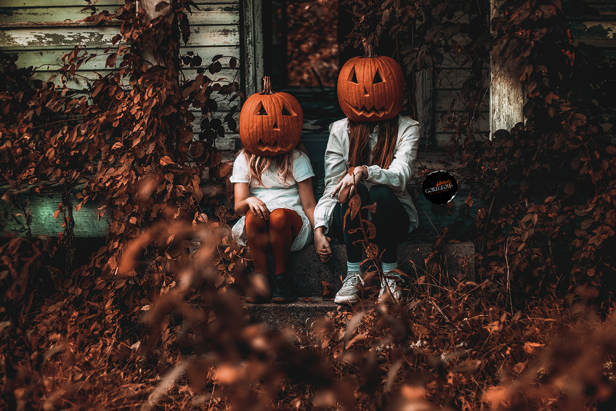 Two children sitting on steps in front of an old house, both wearing pumpkin masks as headwear, surrounded by autumn leaves and foliage.