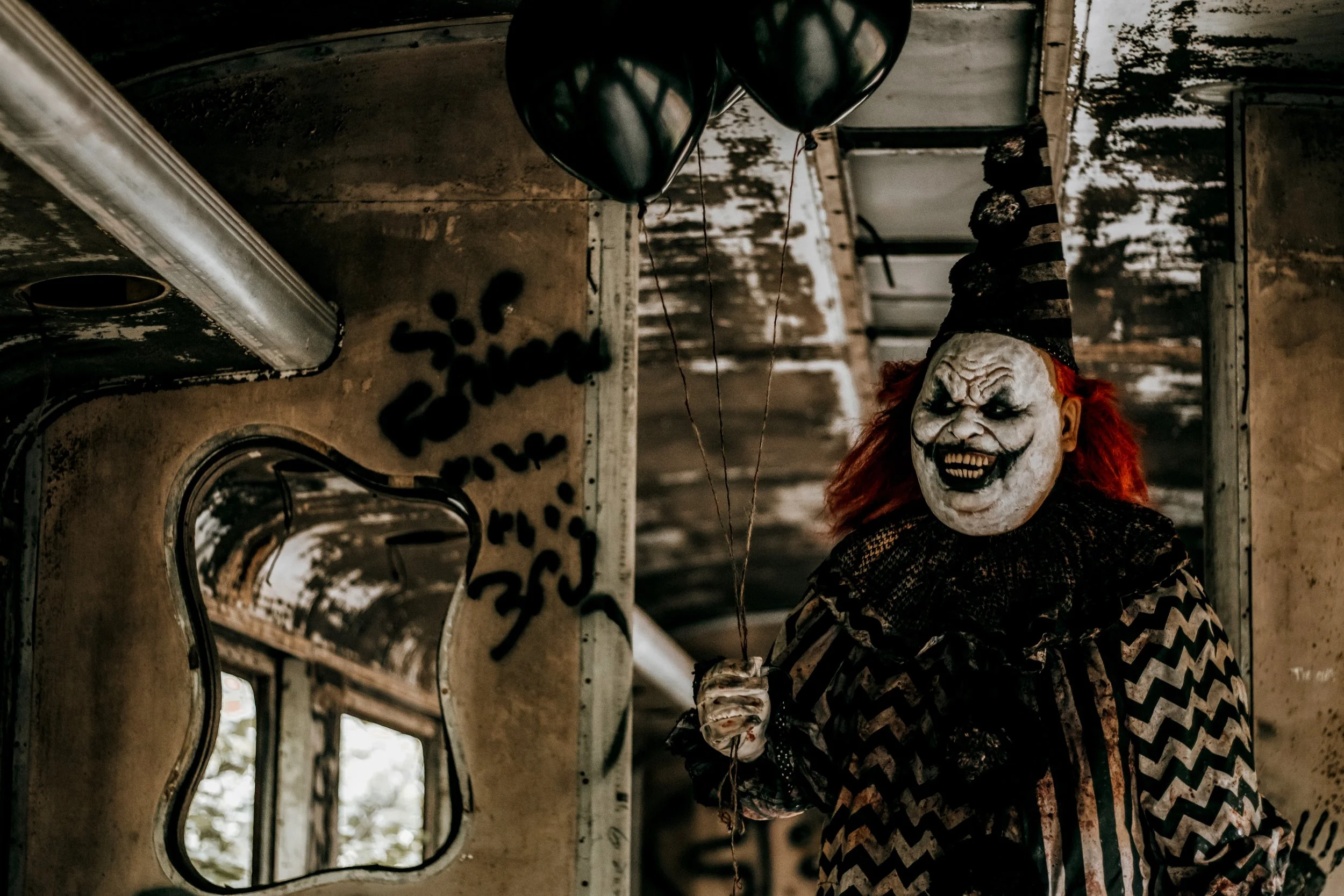 A person dressed as a creepy clown with a white painted face, red hair, and a black hat, standing inside an abandoned, graffiti-covered trailer with dark, eerie lighting.