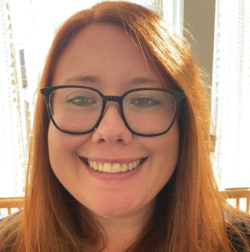 A woman with reddish hair wearing glasses and smiling, indoors near a window with sunlight.