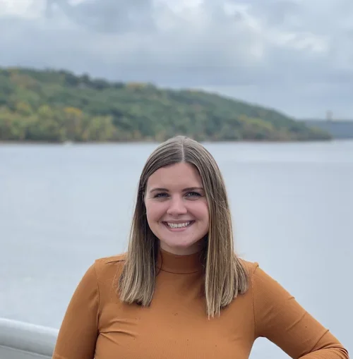 A young woman with shoulder-length hair smiling outdoors near a body of water, with a hilly landscape in the background.