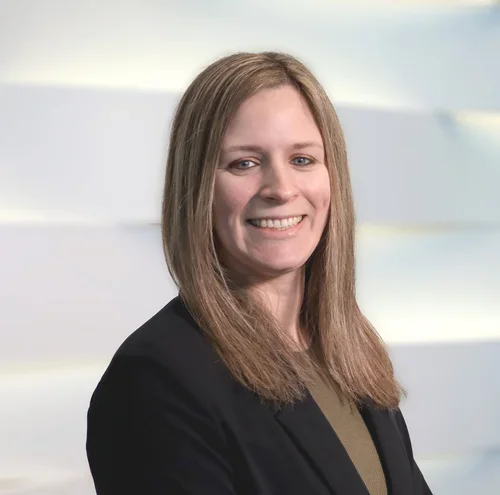 A woman with shoulder-length light brown hair, smiling, wearing a dark blazer and olive green top, standing in front of a light background.