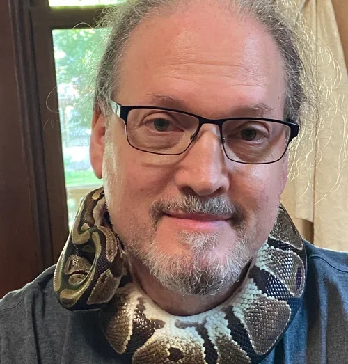 A man with glasses and gray hair is smiling with a snake around his neck.