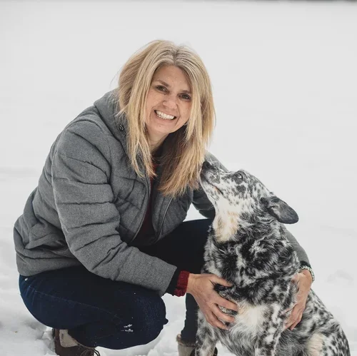 A woman in a gray jacket crouching in snow, smiling, with a merle Australian Shepherd dog in front of her.