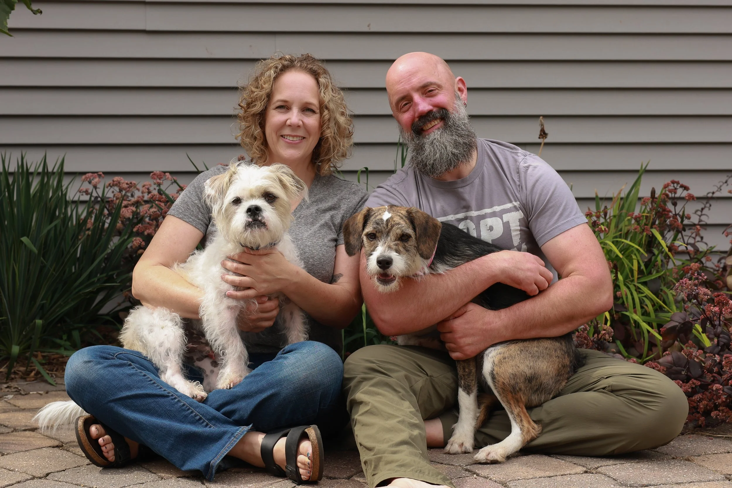 A smiling couple sitting on the ground in front of a house with three dogs, surrounded by garden plants.