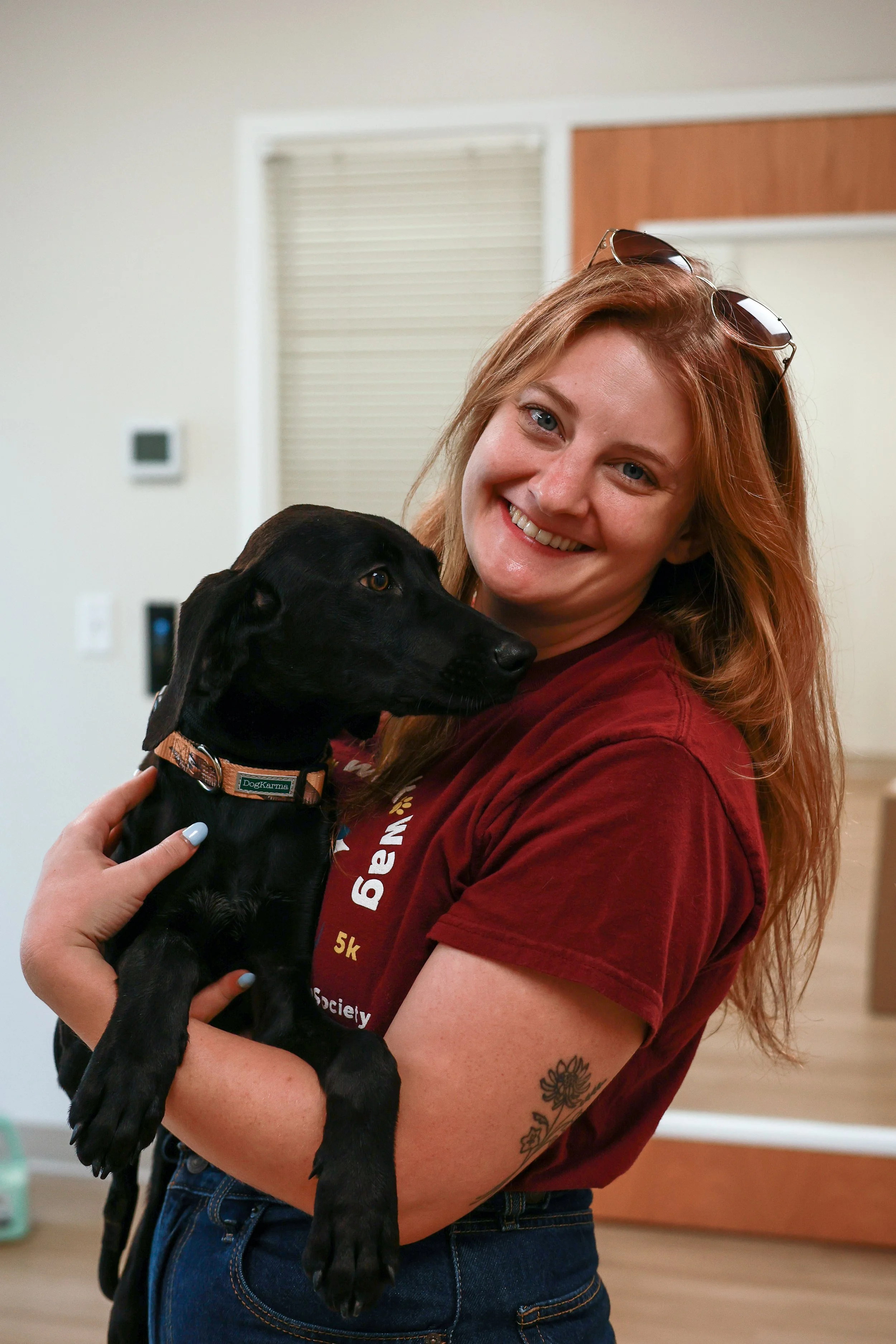 Smiling woman with red hair holding a black puppy with a tan collar indoors.