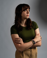 A woman with dark hair wearing a green top and khaki pants, standing with arms crossed against a gray background.
