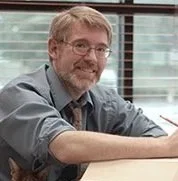 A man with glasses, a beard, and curly hair smiling at a desk in an office setting.