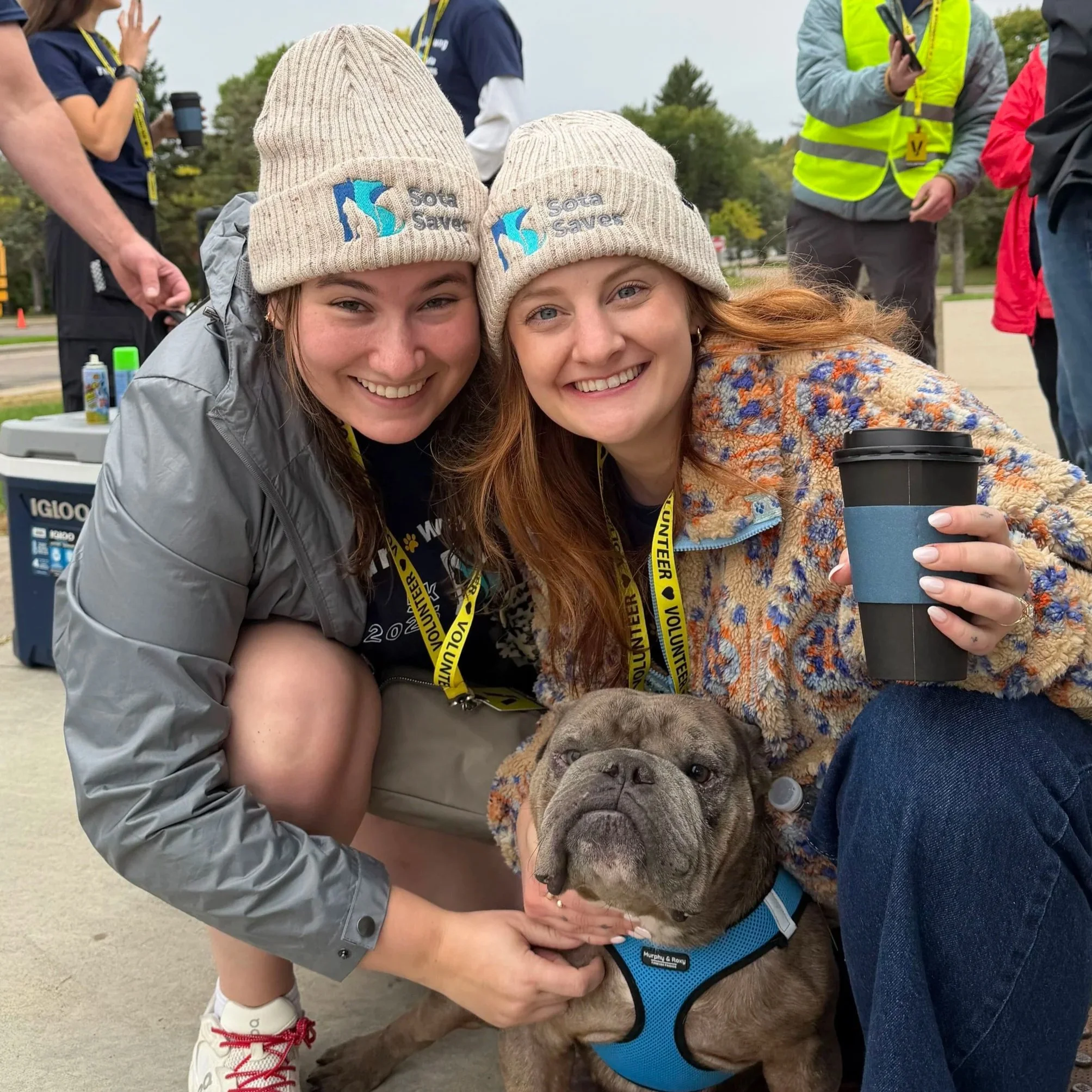 Two smiling women wearing beige charity beanies with blue and gray logos, one is holding a large dog with a blue harness, at an outdoor event with volunteers and trees in the background.