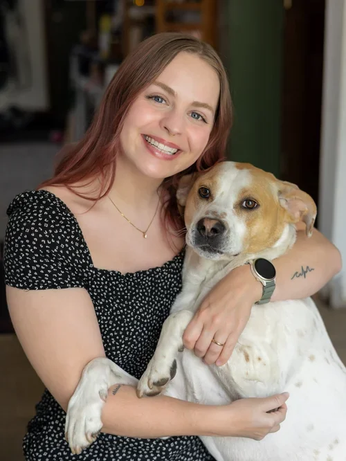 A young woman with long reddish-brown hair smiling and holding a white and brown dog in a cozy indoor setting.