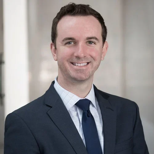 A man in a suit smiling with a blurred office background.