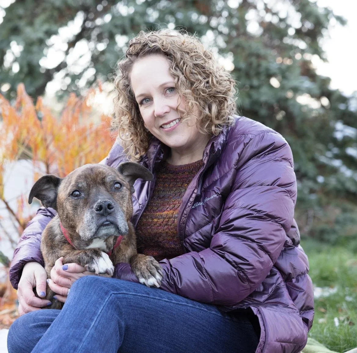 A woman with curly hair smiling and sitting outdoors on grass, holding her brindle dog with a pink collar, surrounded by trees and autumn leaves.