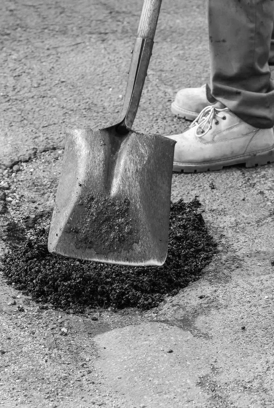 A person wearing work boots is using a shovel to move dark soil or asphalt on a rough ground surface.
