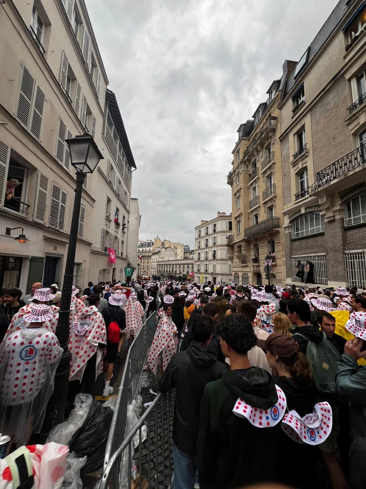 Crowd of people dressed in humorous polka dot outfits gathered on a city street, lined with tall Parisian-style buildings, under overcast skies.