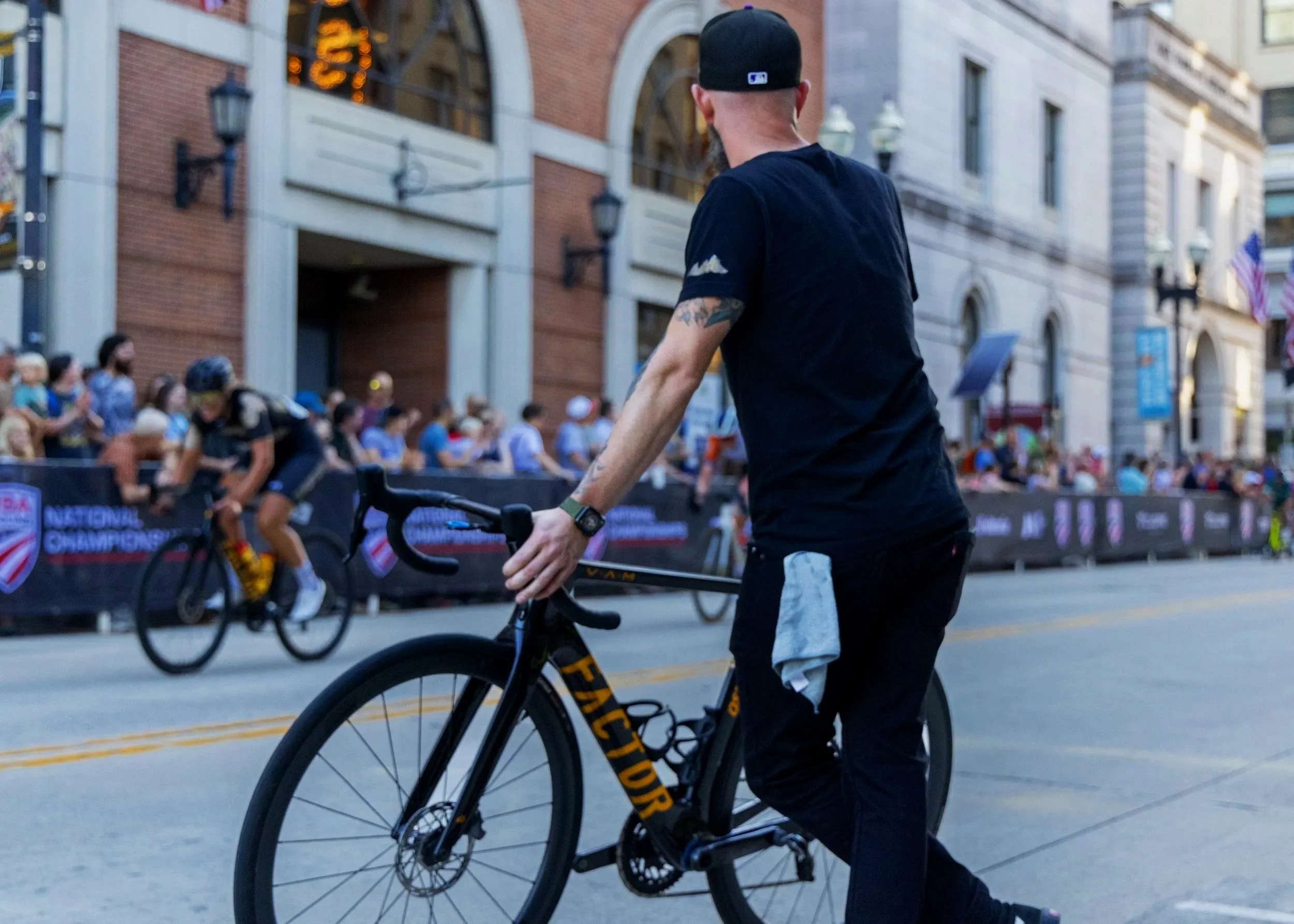 A person wearing a black cap and black t-shirt riding a black and yellow bicycle on a city street with a crowd of people in the background and historic buildings.