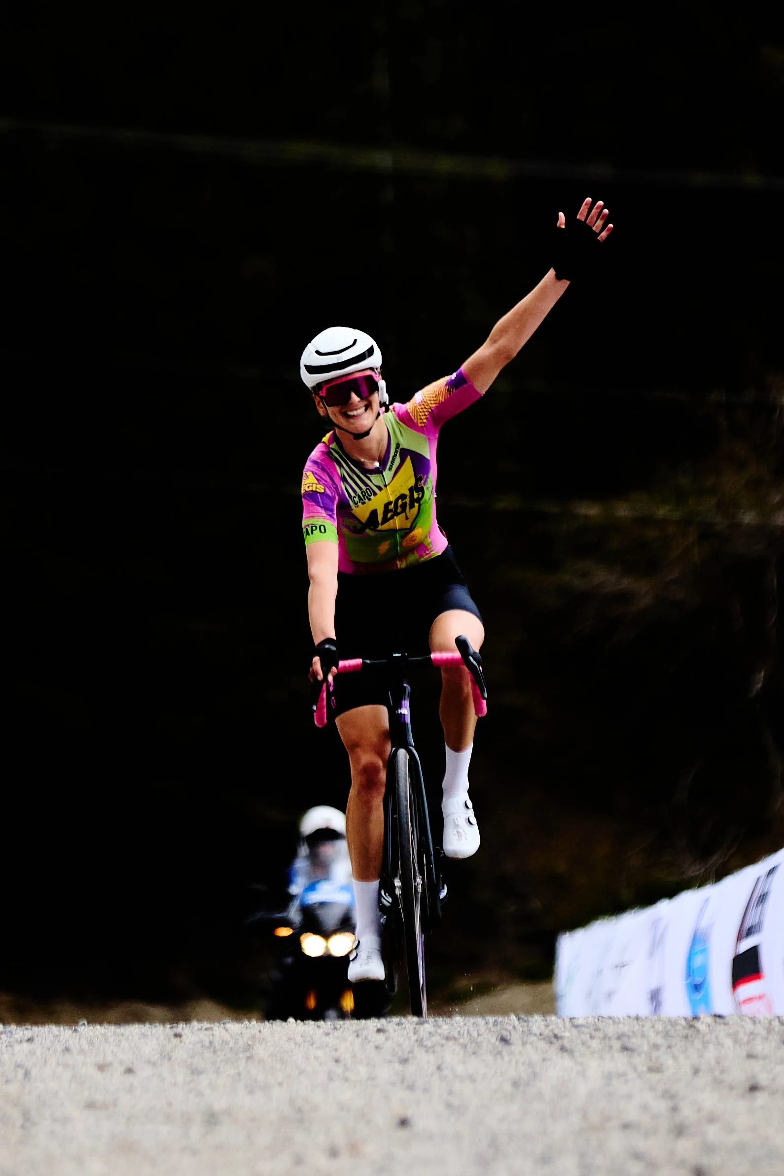 A person riding a bike wearing a purple and yellow jersey, a white helmet, and sunglasses, with one arm raised in a celebratory gesture. The background is dark, with some spectators and a banner visible.