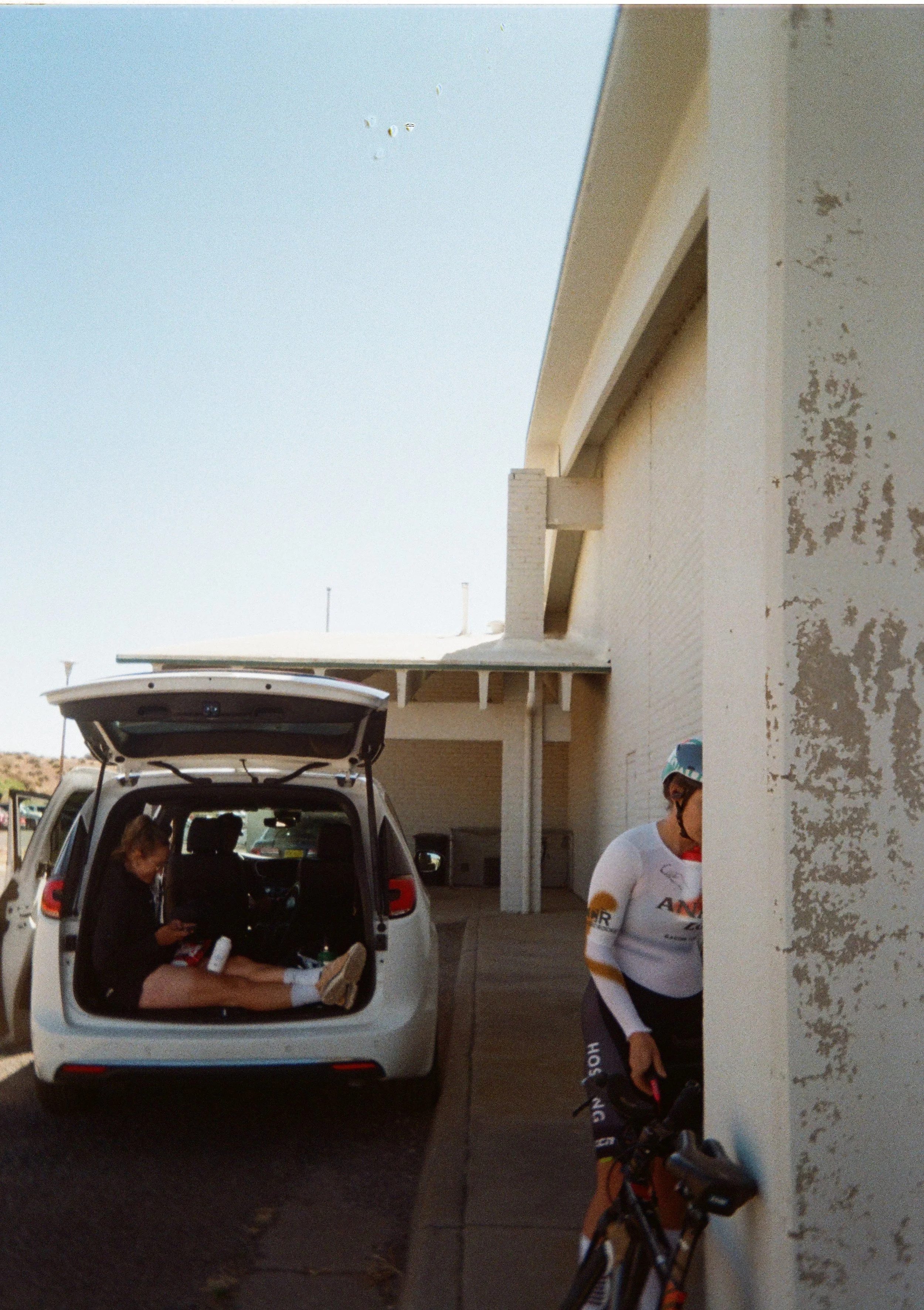 A woman sitting in the open trunk of a white SUV, looking at her phone, with two people standing nearby. One person is wearing a white cycling jersey and holding a scooter. A bicycle is leaning against the wall.