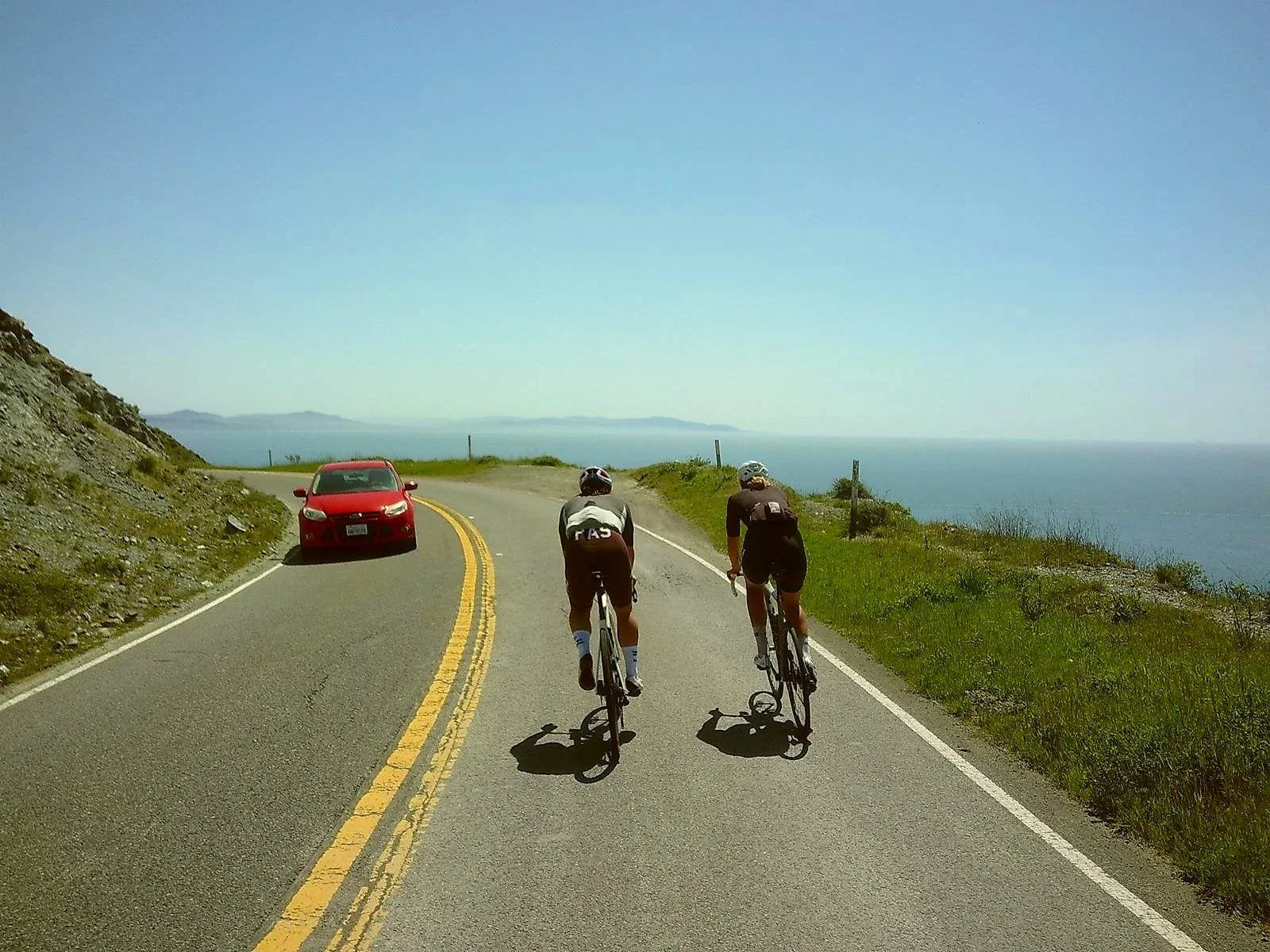 Two cyclists riding on a coastal road with a red car ahead and ocean view to the right.