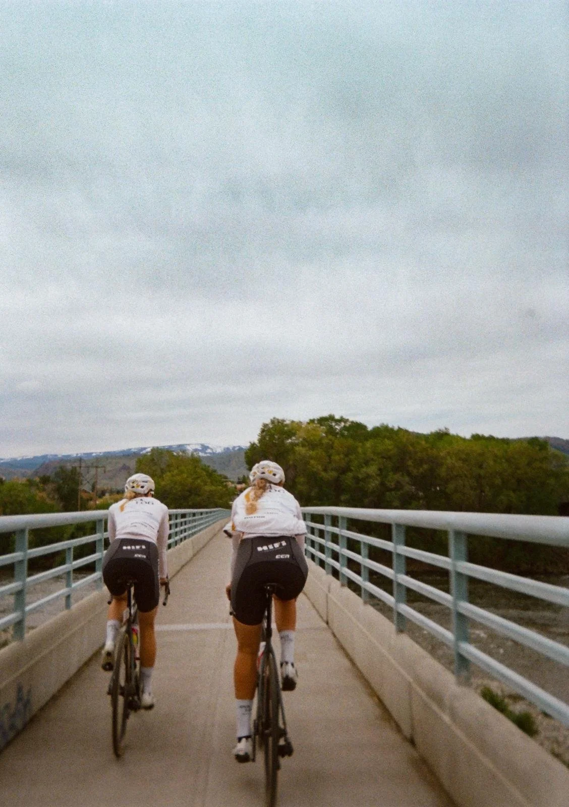Two cyclists riding on a bridge with metal railings, trees, and mountains in the background under a cloudy sky.