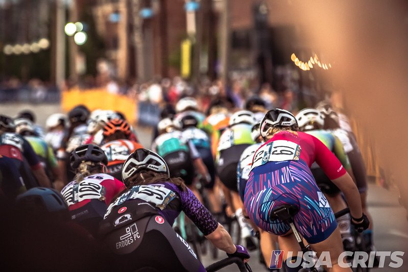 Cyclists participating in a competitive race, riding closely together on a city street, with spectators and buildings in the background.