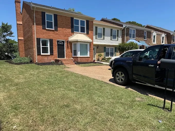 A brick two-story house with black window shutters, a white front door, and an upper porch, alongside a driveway with a black pickup truck, in a suburban neighborhood with well-maintained lawns.