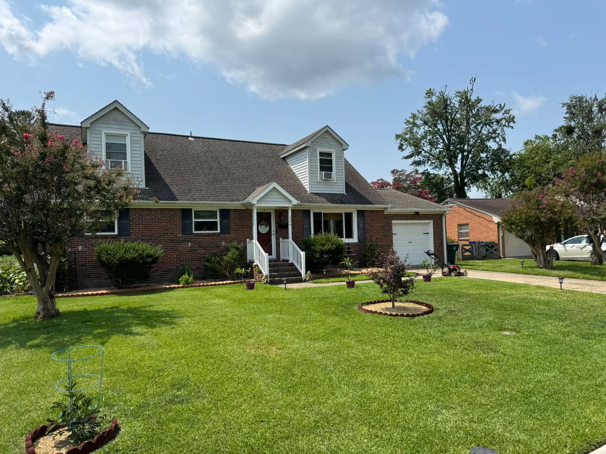Front yard of a brick house with white siding, black shutters, a small porch with white railing, and a driveway with a white car. The yard has lush green grass, trees with blossoms, and a small garden bed.